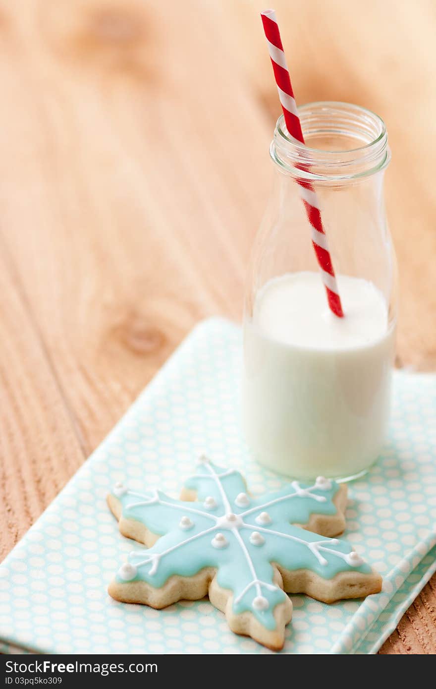 One blue snowflake sugar cookie rests on blue napkin next to a small farm bottle of milk with a red and white striped straw. One blue snowflake sugar cookie rests on blue napkin next to a small farm bottle of milk with a red and white striped straw.