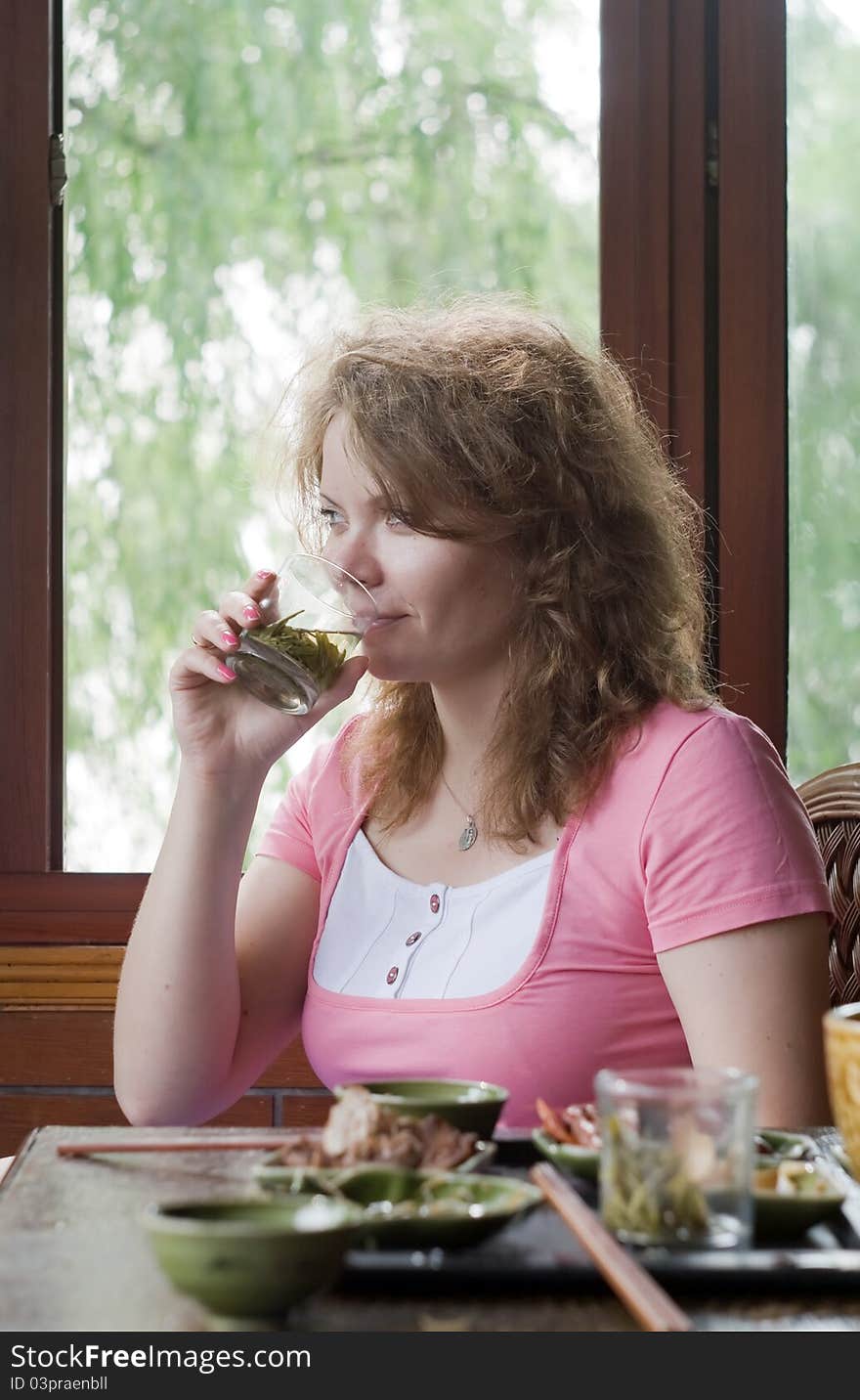 Portrait image of woman which is drinking the green tea opposite the window with beautiful view on the nature. On the foreground is Chinese food on the table. Young woman is smiling. Portrait image of woman which is drinking the green tea opposite the window with beautiful view on the nature. On the foreground is Chinese food on the table. Young woman is smiling.