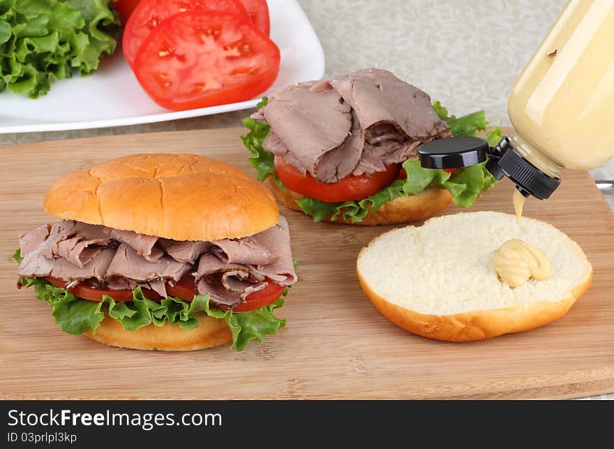 Roast beef sandwich with lettuce and tomato and pouring mustard onto a bun