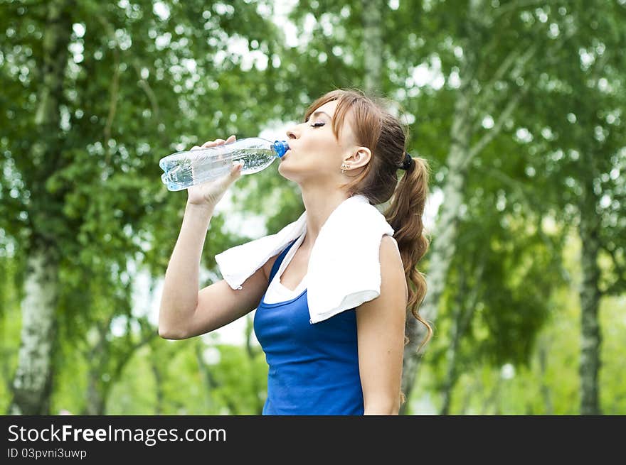 Young woman drinking water after exercise, summer park background