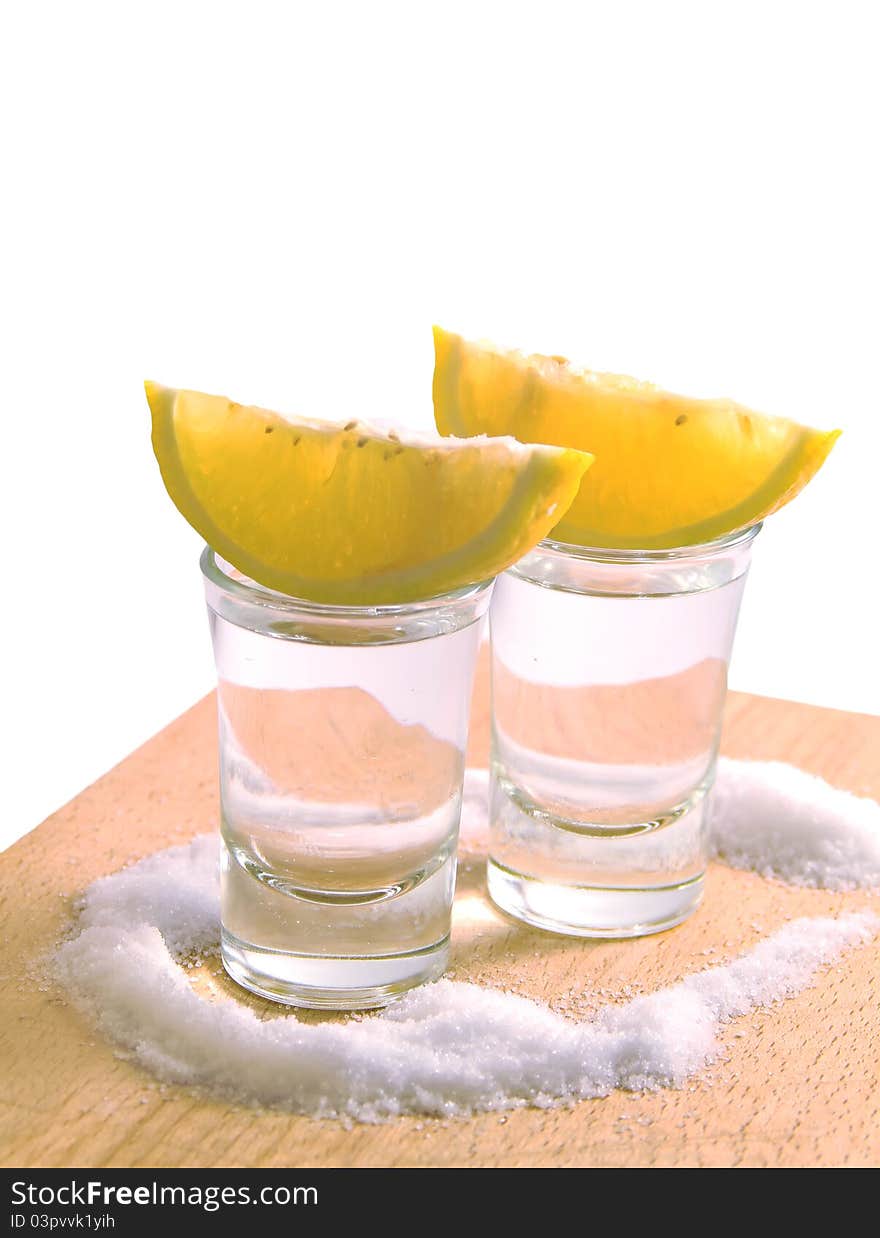 Two glasses filled with tequila, with slices of lemon on the top of it, all surronded with salt, isolated in white. Two glasses filled with tequila, with slices of lemon on the top of it, all surronded with salt, isolated in white