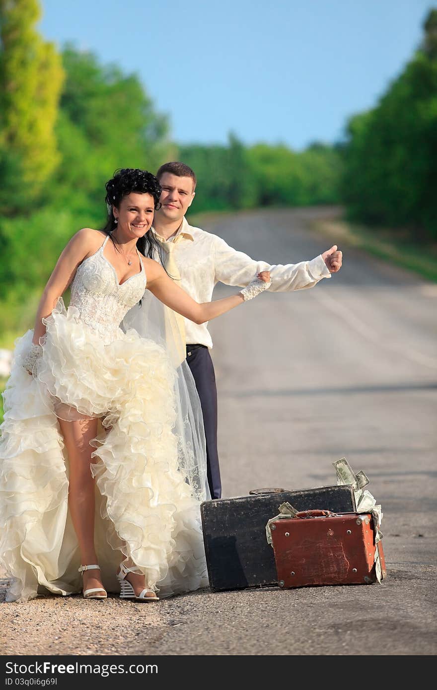 Young newlywed couple with suitcases of cash  hitchhiking on a road. Young newlywed couple with suitcases of cash  hitchhiking on a road.