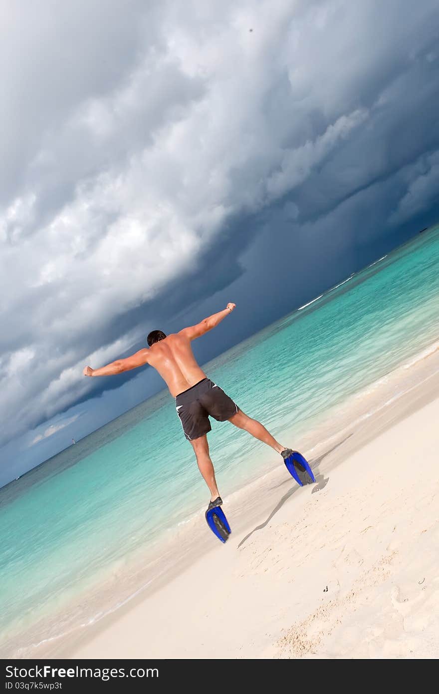 Tanned man jump in blue flippers on sand beach and ocean under gloomy sky