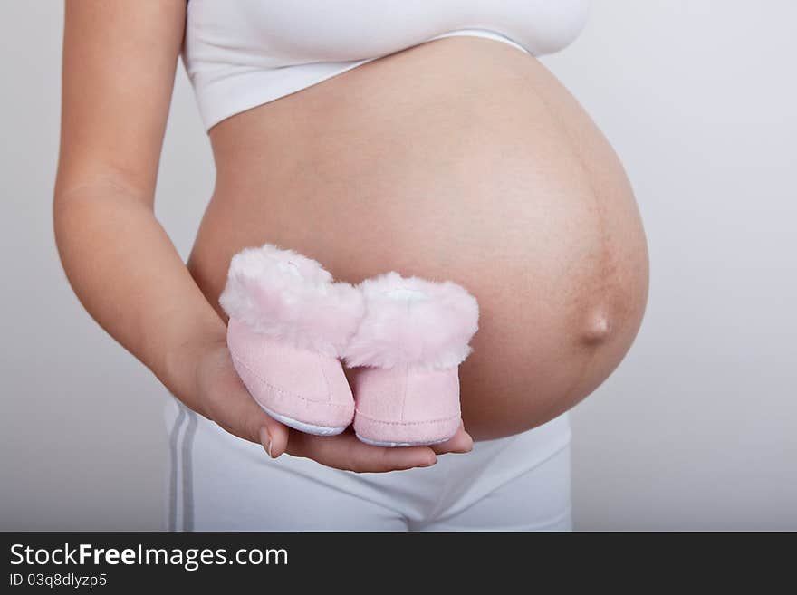 Young european pregnant woman hold in hand pink baby boots on grey background. Young european pregnant woman hold in hand pink baby boots on grey background