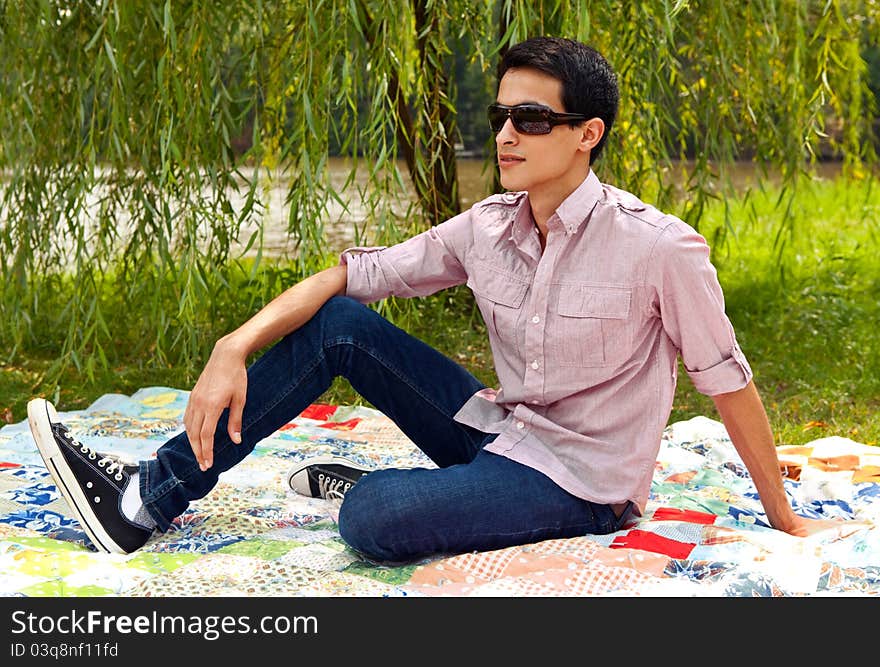 A portrait of a handsome young man, sitting outside on a picnic blanket on a sunny day, with a willow tree behind him. A portrait of a handsome young man, sitting outside on a picnic blanket on a sunny day, with a willow tree behind him