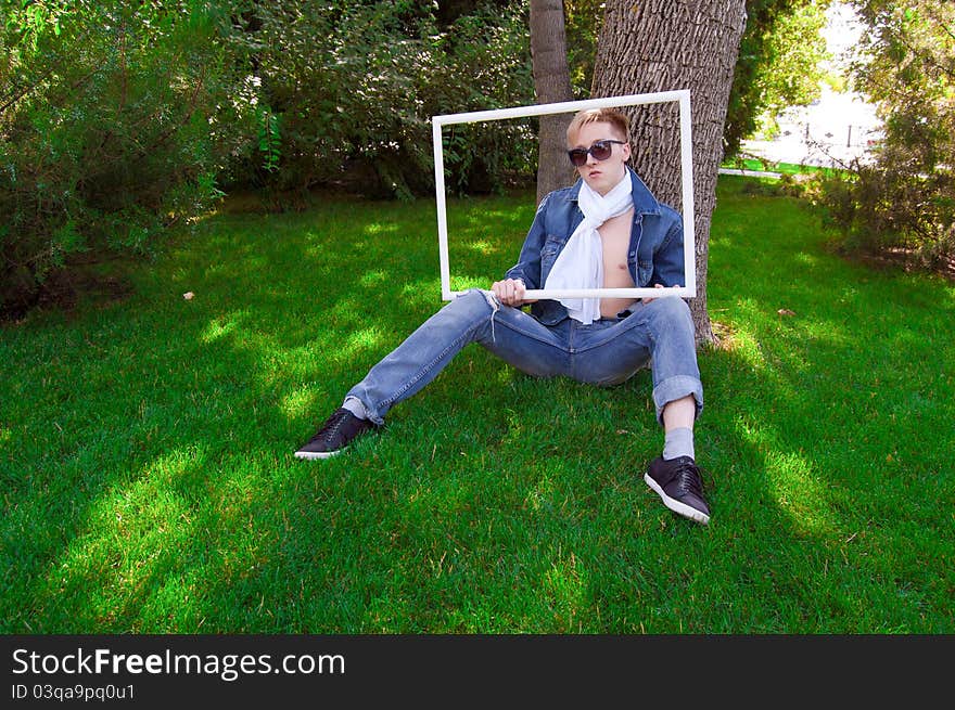 Young human is siting on the green grass with white frame in his hand, he has blue goggles and jean jacket and white scarf he is funny. Young human is siting on the green grass with white frame in his hand, he has blue goggles and jean jacket and white scarf he is funny