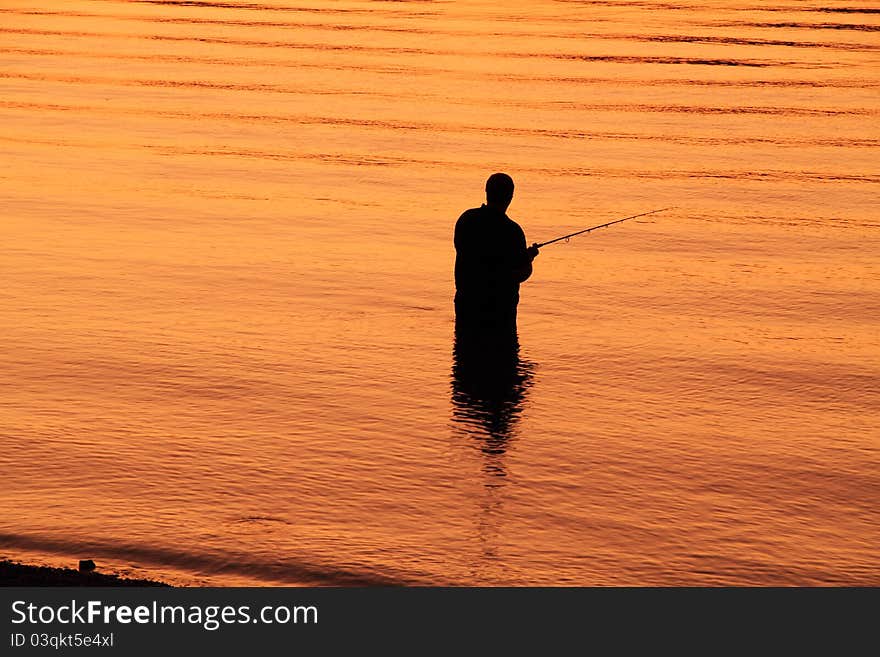 A lone fisherman is silhouetted against the water which is golden orange because of the setting sun. A lone fisherman is silhouetted against the water which is golden orange because of the setting sun.