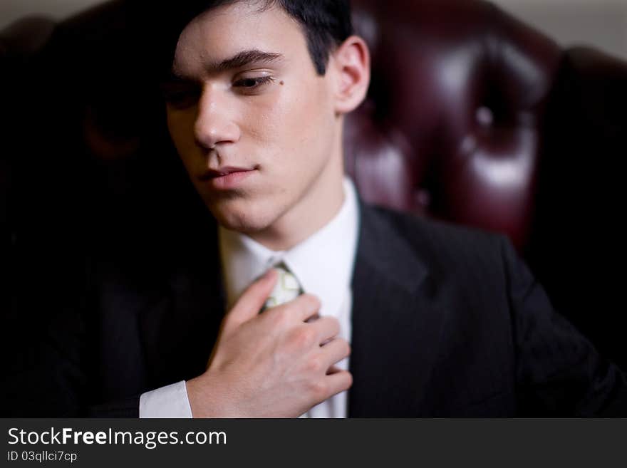 Portrait of a pensive young man dressed in a business suit, touching his tie, seated in a plush leather chair and looking down camera left. Portrait of a pensive young man dressed in a business suit, touching his tie, seated in a plush leather chair and looking down camera left.