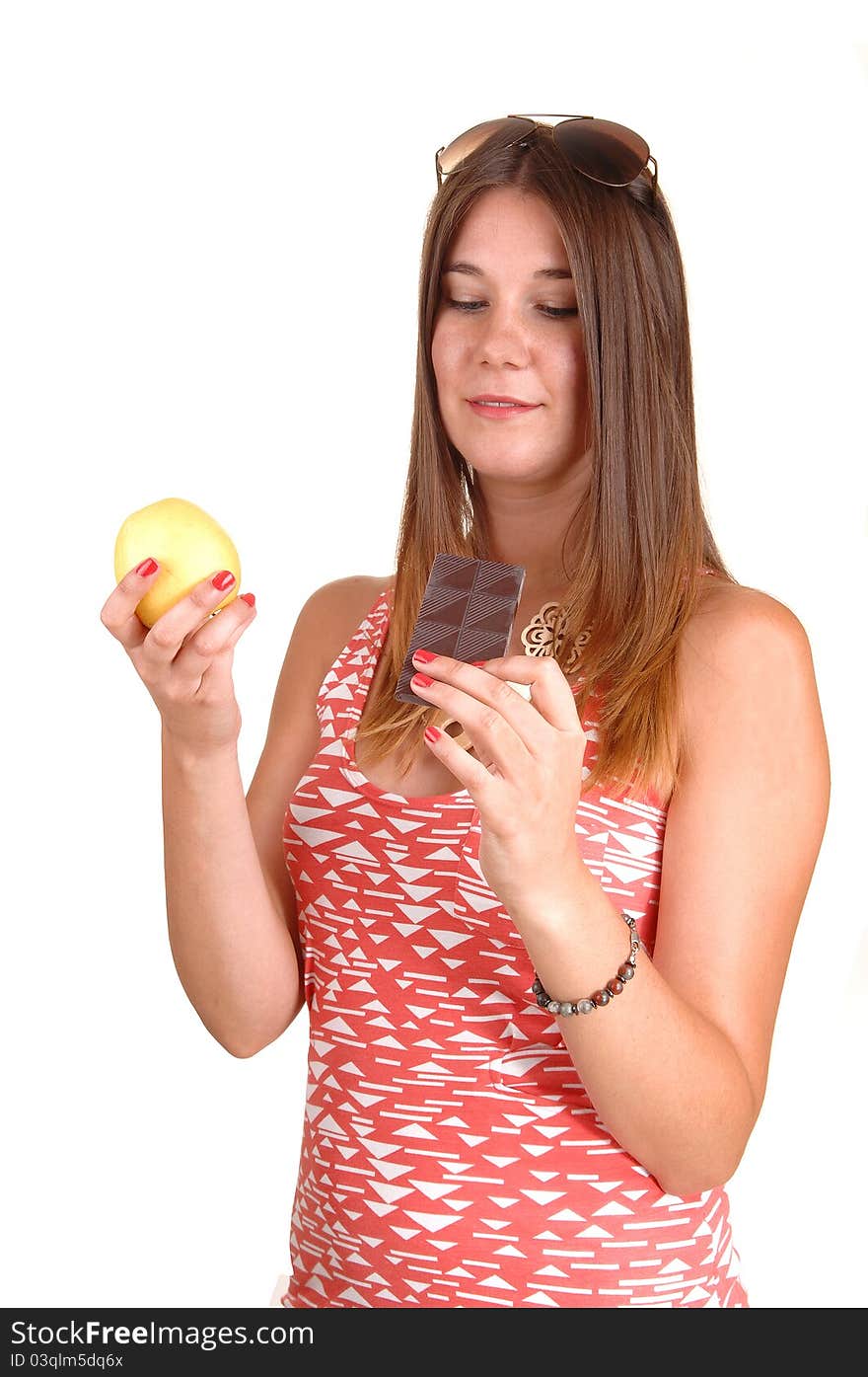 A young woman in a pink dress and sunglasses on her head can not decide to eat an apple or chocolate, in white background. A young woman in a pink dress and sunglasses on her head can not decide to eat an apple or chocolate, in white background.