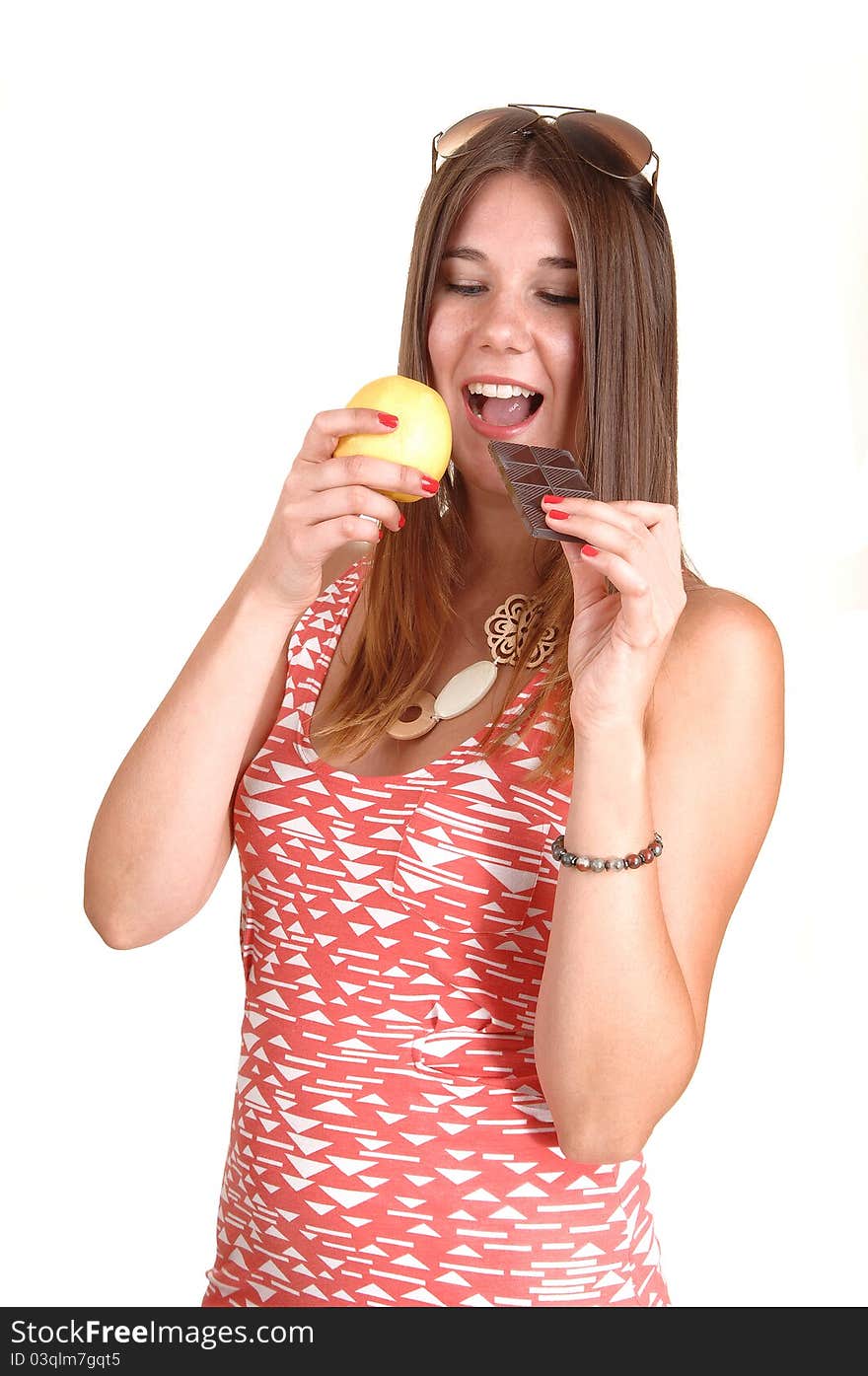 A young woman in a pink dress and sunglasses on her head can not decide to eat an apple or chocolate, in white background. A young woman in a pink dress and sunglasses on her head can not decide to eat an apple or chocolate, in white background.