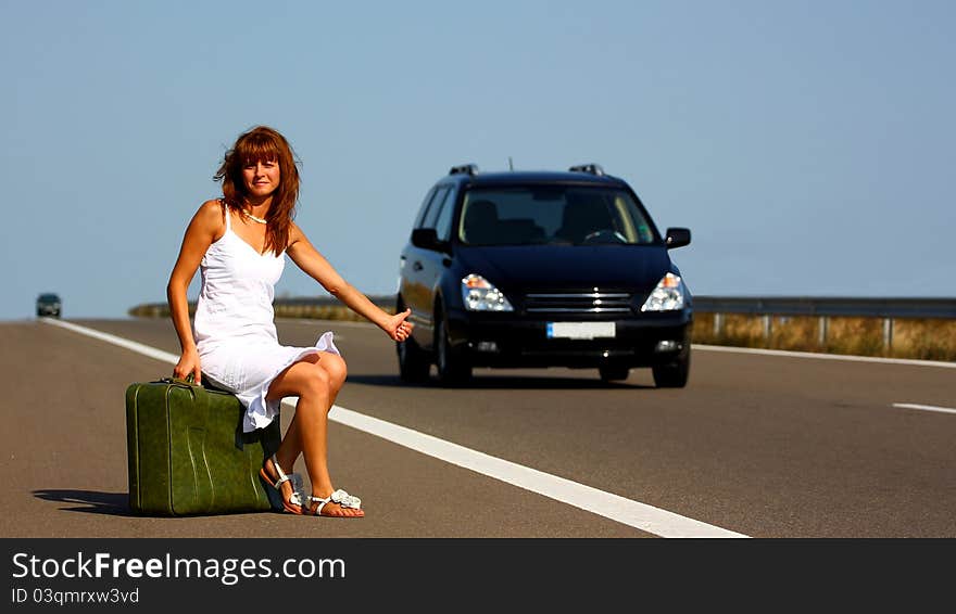 Woman hitchhiking on a highway sitting on a green briefcase