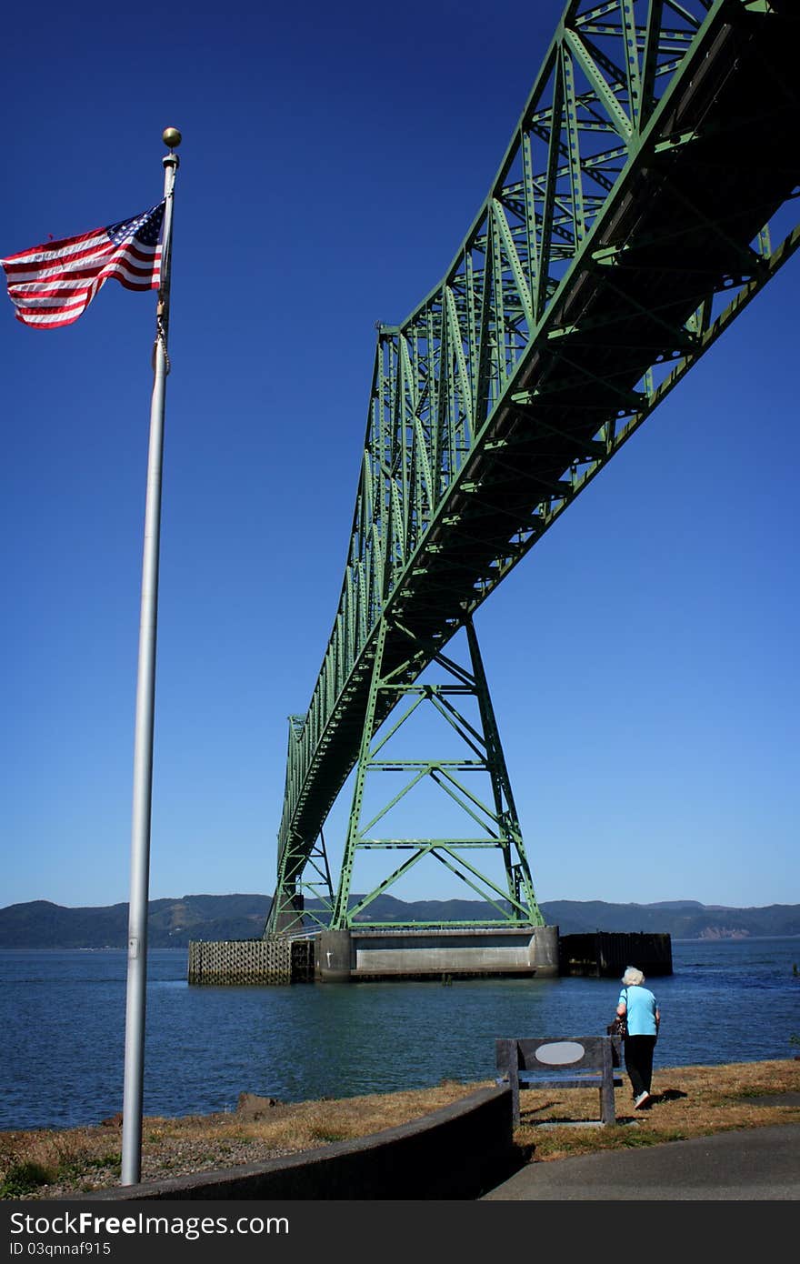 A lady stands under and looks at the historic 4.2 mile long Astoria Megler Bridge which goes over the mouth of the Columbia River in the Pacific Northwest, viewing from the Astoria Oregon side with an American Flag flying. Clear sunny blue skies. 2000 cargo ships a year pass underneath it. A lady stands under and looks at the historic 4.2 mile long Astoria Megler Bridge which goes over the mouth of the Columbia River in the Pacific Northwest, viewing from the Astoria Oregon side with an American Flag flying. Clear sunny blue skies. 2000 cargo ships a year pass underneath it.