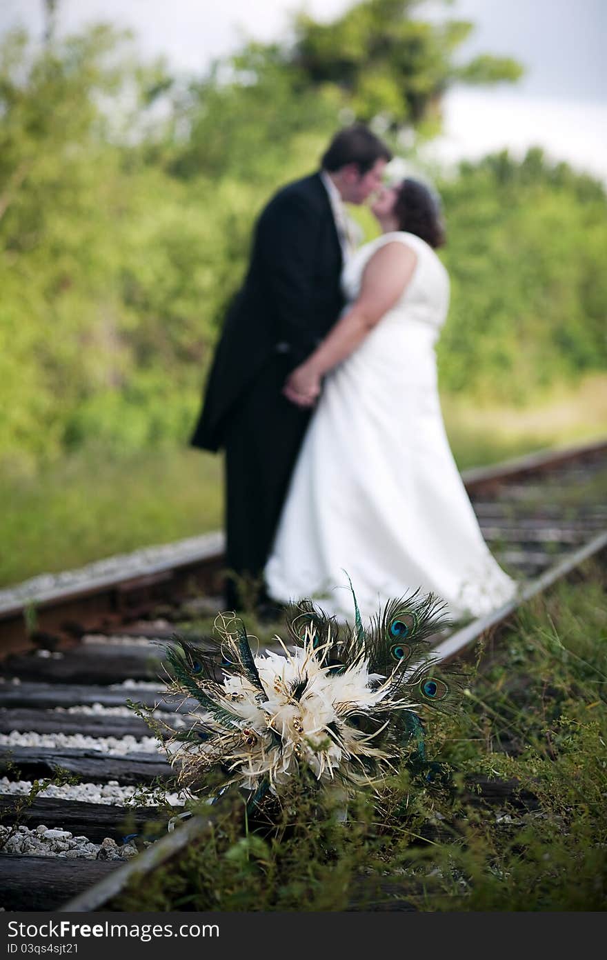 A feather wedding bouquet on railroad tracks with wedding couple standing holding hands behind and out of focus. A feather wedding bouquet on railroad tracks with wedding couple standing holding hands behind and out of focus.