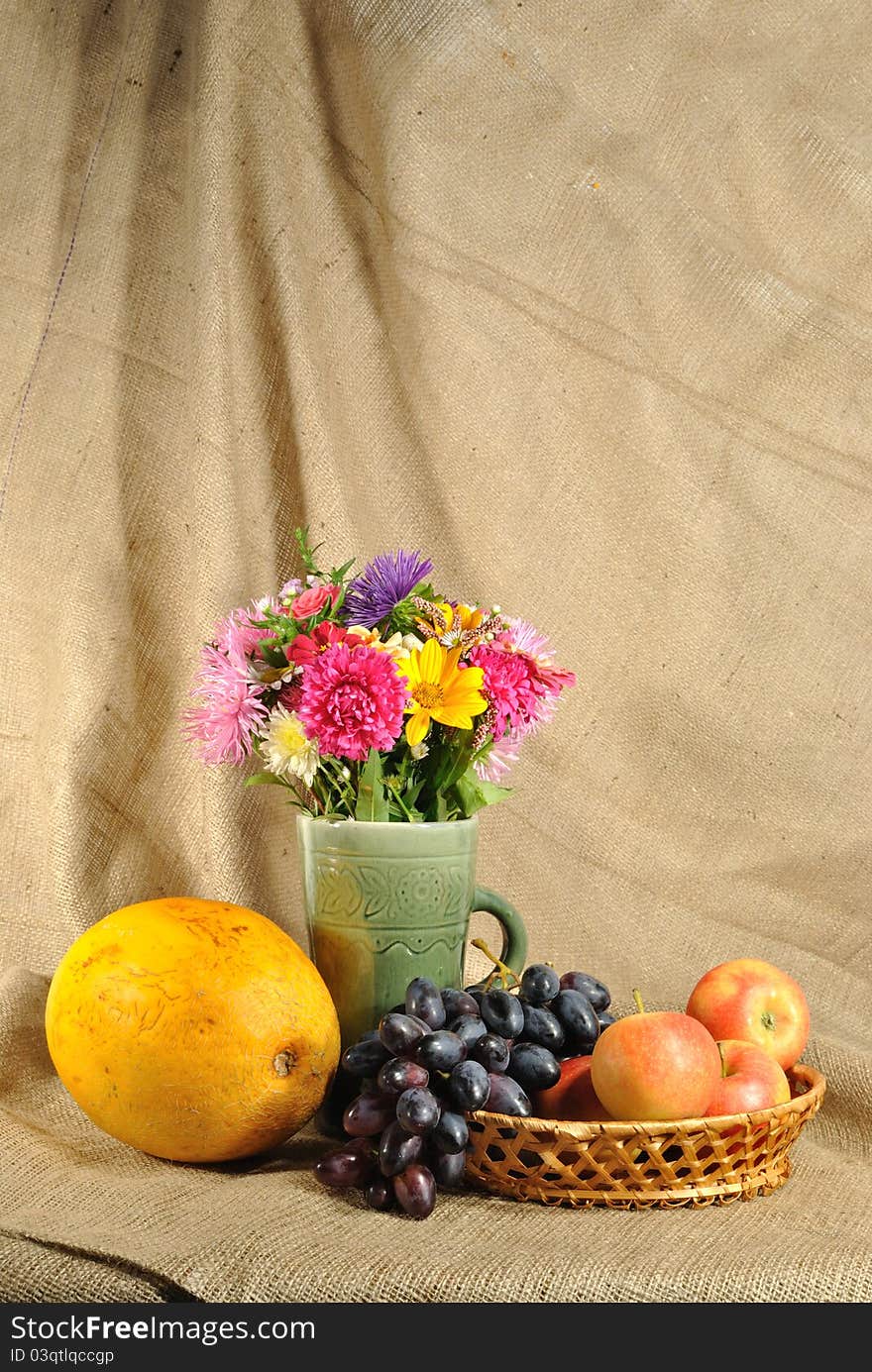 The autumn topic. In the foreground is bouquet of wild and cultivated flowers, bunch of black grapes and melon. They are on the table covered by rough stuff. This staff creates background. The autumn topic. In the foreground is bouquet of wild and cultivated flowers, bunch of black grapes and melon. They are on the table covered by rough stuff. This staff creates background.