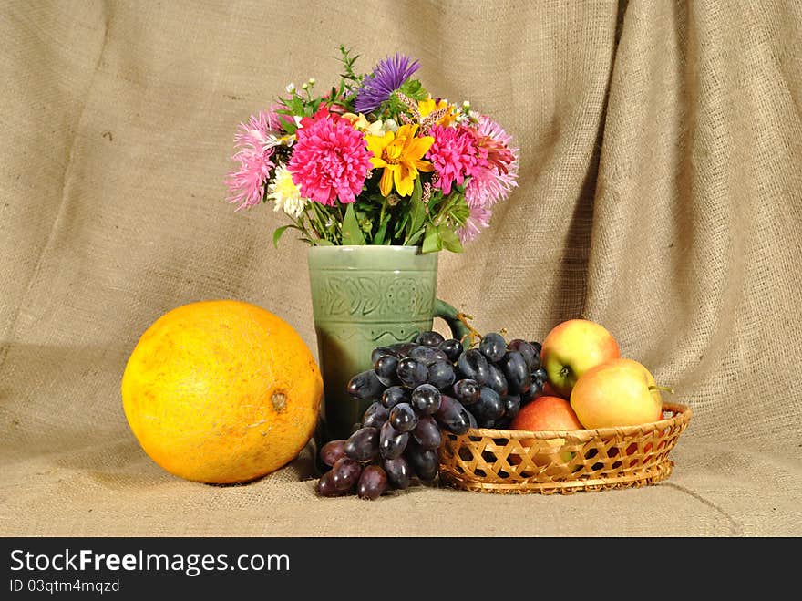 The autumn topic. In the foreground is bouquet of wild and cultivated flowers, bunch of black grapes and melon. They are on the table covered by rough stuff. This staff creates background. The autumn topic. In the foreground is bouquet of wild and cultivated flowers, bunch of black grapes and melon. They are on the table covered by rough stuff. This staff creates background.