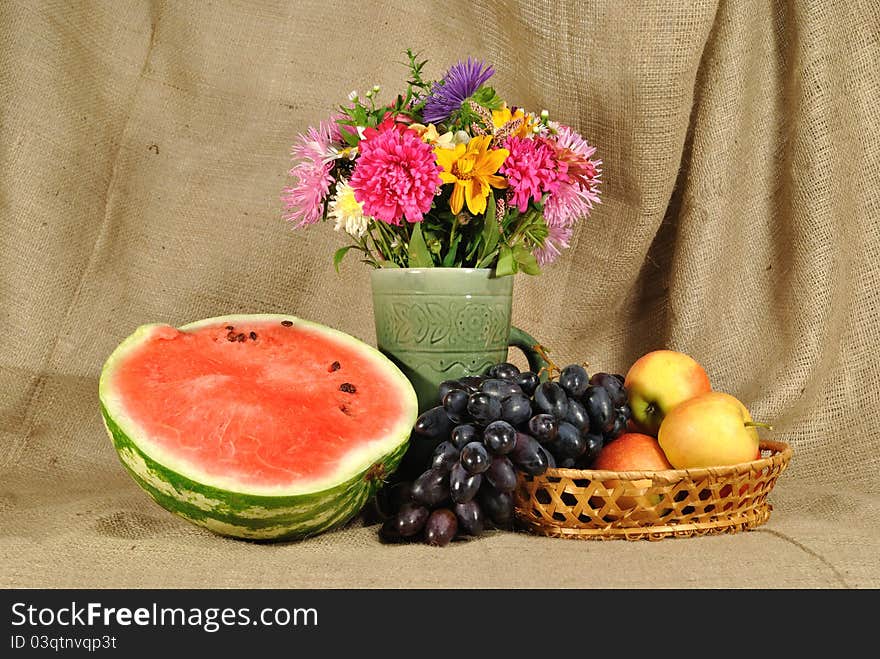 The autumn topic. In the foreground is bouquet of wild and cultivated flowers, bunch of black grapes and melon. They are on the table covered by rough stuff. This staff creates background. The autumn topic. In the foreground is bouquet of wild and cultivated flowers, bunch of black grapes and melon. They are on the table covered by rough stuff. This staff creates background.