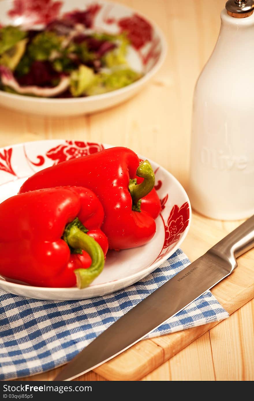 Two red peppers on a table with silver knife and salad in the background.