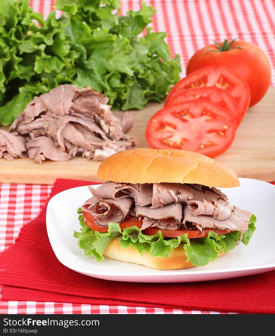 Roast beef sandwich with lettuce and tomato on a plate with ingredients in background. Roast beef sandwich with lettuce and tomato on a plate with ingredients in background