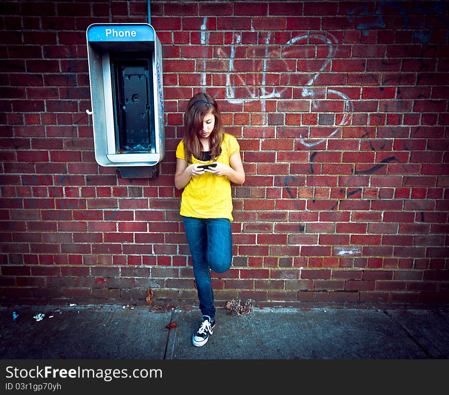 A cute teenage girl standing next to an obsolete payphone while texting on a cell phone against a grungy urban wall. A cute teenage girl standing next to an obsolete payphone while texting on a cell phone against a grungy urban wall