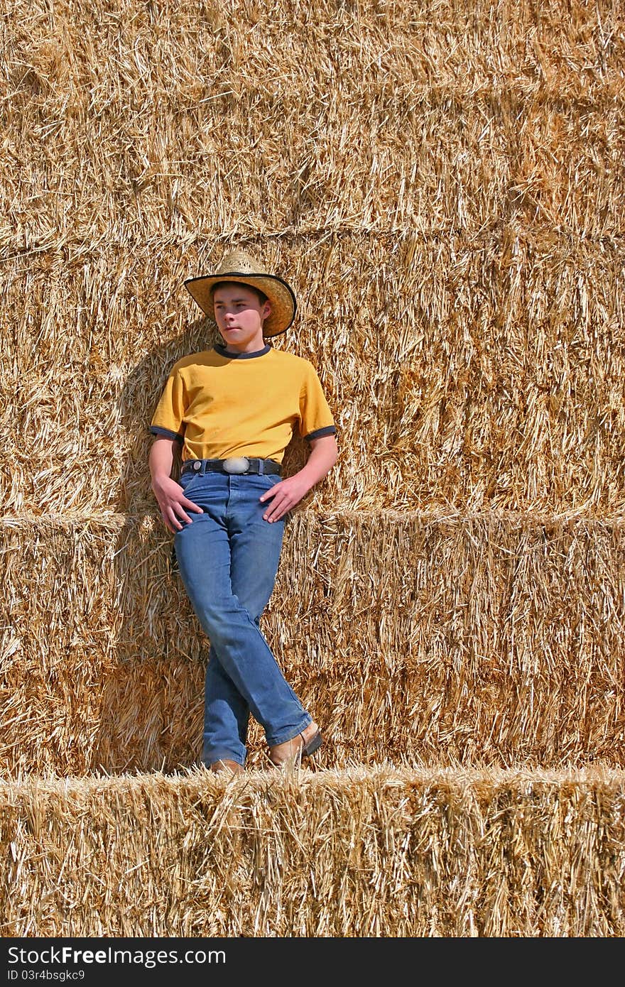 Male cowboy standing on straw stack with cowboy hat wearing yellow shirt and blue jeans in the sun looking off in the distance. Male cowboy standing on straw stack with cowboy hat wearing yellow shirt and blue jeans in the sun looking off in the distance