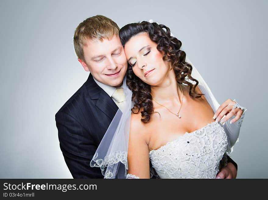 Studio portrait of young beautiful happy just married bride and groom with closed eyes embracing on blue background