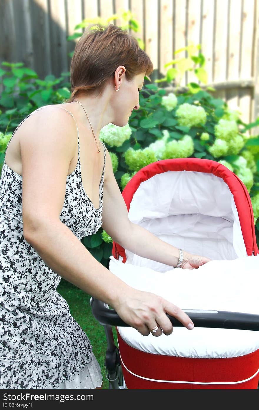 Beautiful young happy smiling woman looking in red baby stroller near the flowers. Beautiful young happy smiling woman looking in red baby stroller near the flowers