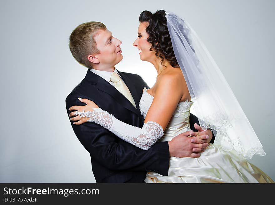 Studio portrait of young elegant enamored just married bride and groom looking to each other and embracing on grey background