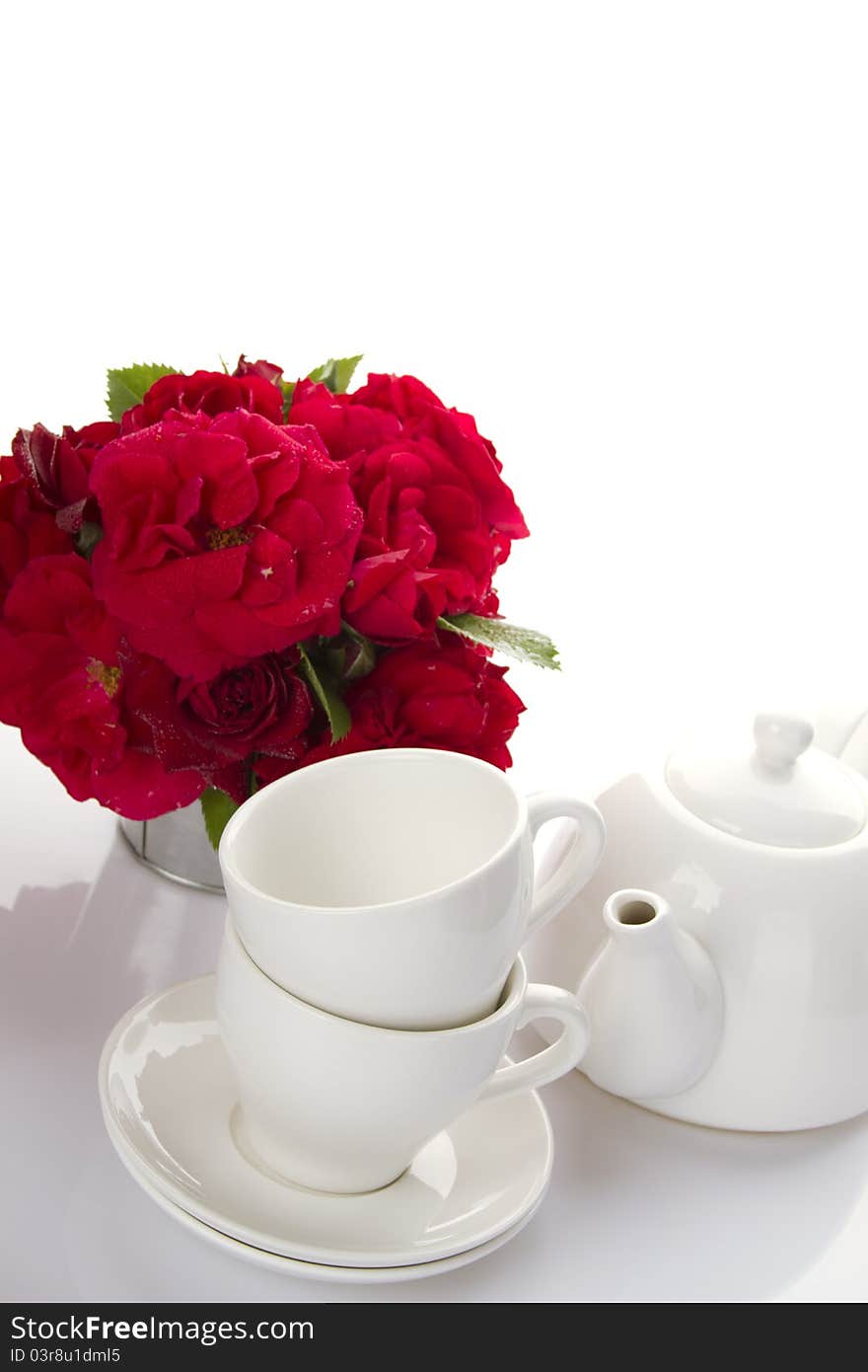 White crockery for tea kettle, two cups and saucers and a bouquet of red roses in a small iron bucket. White crockery for tea kettle, two cups and saucers and a bouquet of red roses in a small iron bucket