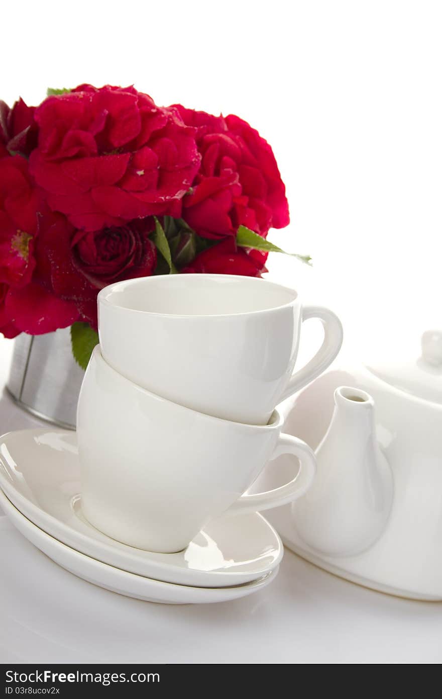 White crockery for tea kettle, two cups and saucers and a bouquet of red roses in a small iron bucket. White crockery for tea kettle, two cups and saucers and a bouquet of red roses in a small iron bucket