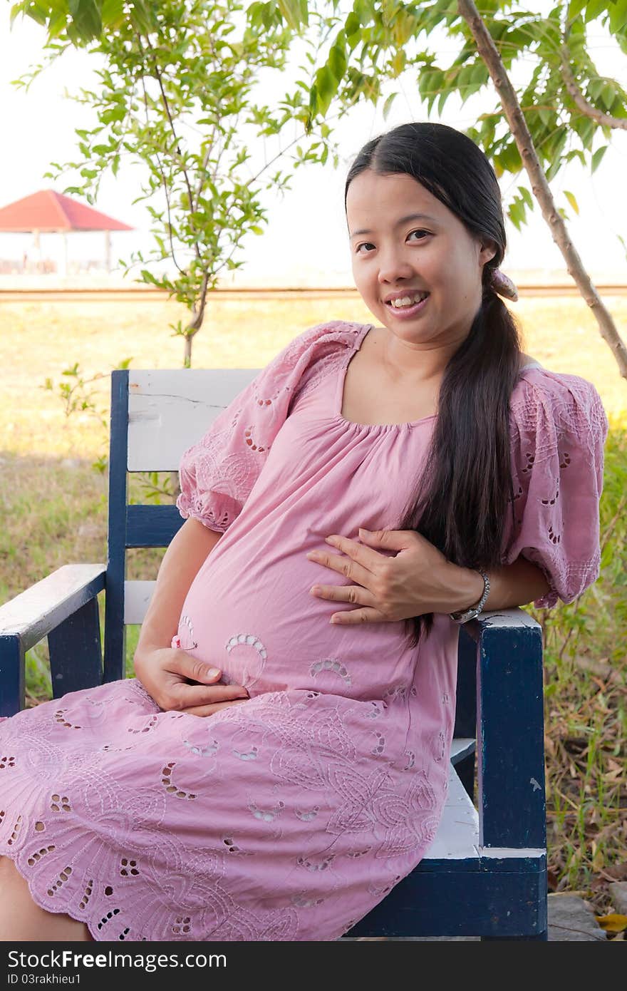 Asian pregnant woman in pink dress smiling in park. Asian pregnant woman in pink dress smiling in park