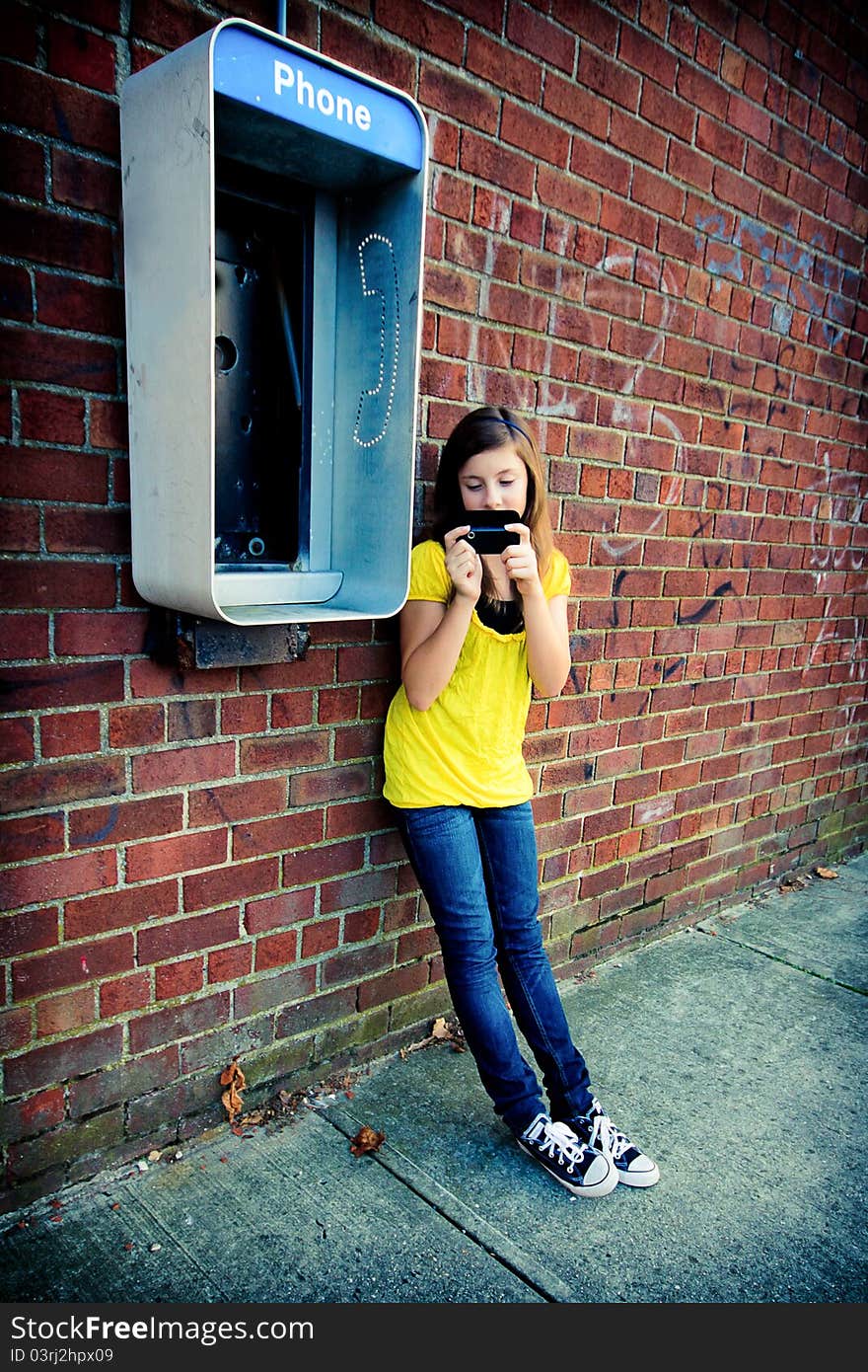 A cute teenage girl texting on her cell phone against a grungy urban wall with an obsolete payphone. A cute teenage girl texting on her cell phone against a grungy urban wall with an obsolete payphone