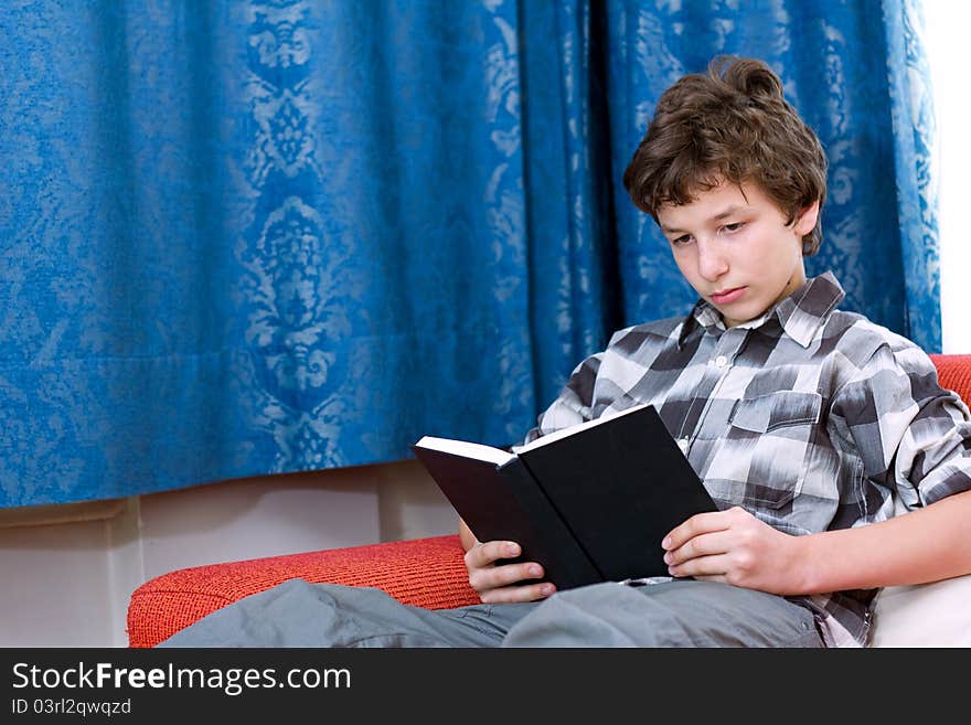 A pre-teen boy in a checkered shirt reading a blank black book on an orange couch with blue damask curtains in the background. A pre-teen boy in a checkered shirt reading a blank black book on an orange couch with blue damask curtains in the background.