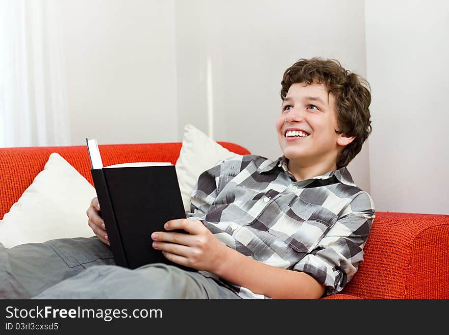 A preteen boy laying back on an orange couch with white pillows as he holds a black book in his hand and looks up with a big smile. A preteen boy laying back on an orange couch with white pillows as he holds a black book in his hand and looks up with a big smile.