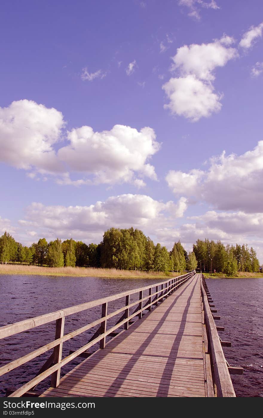 Long wooden footbridge with handrails over the lake and the man walking far away. Forest in the distance and the cloudy sky. Long wooden footbridge with handrails over the lake and the man walking far away. Forest in the distance and the cloudy sky.