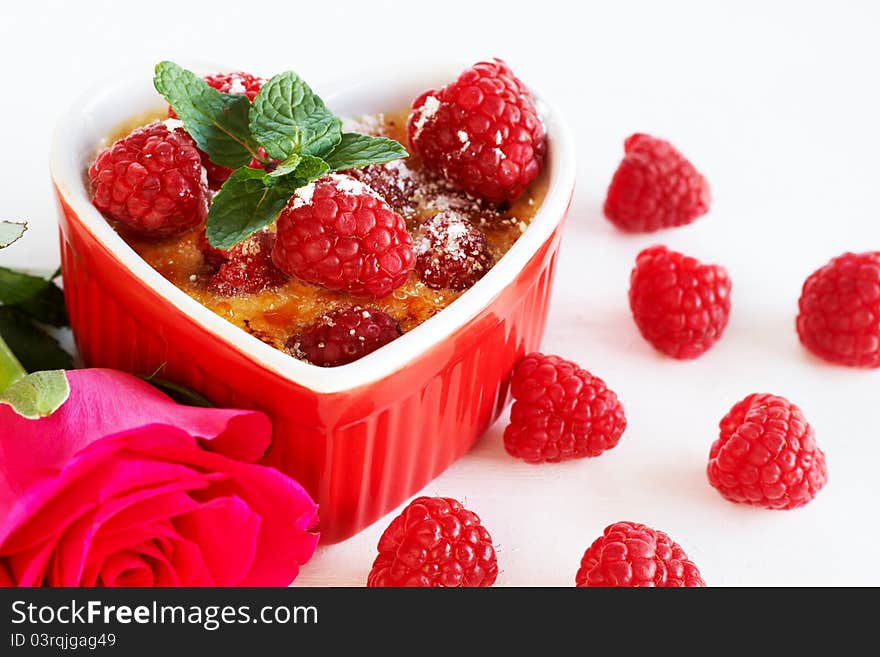 French creme brulee dessert with raspberries and mint covered with caramelized sugar in red heart shaped ramekin on white background