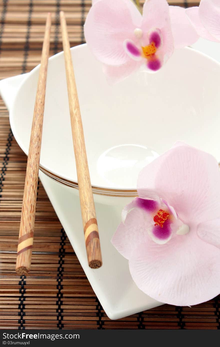 A beautiful Asian place setting including a white rice bowl on a square white plate, wooden chopsticks and orchids on a bamboo mat. A beautiful Asian place setting including a white rice bowl on a square white plate, wooden chopsticks and orchids on a bamboo mat.