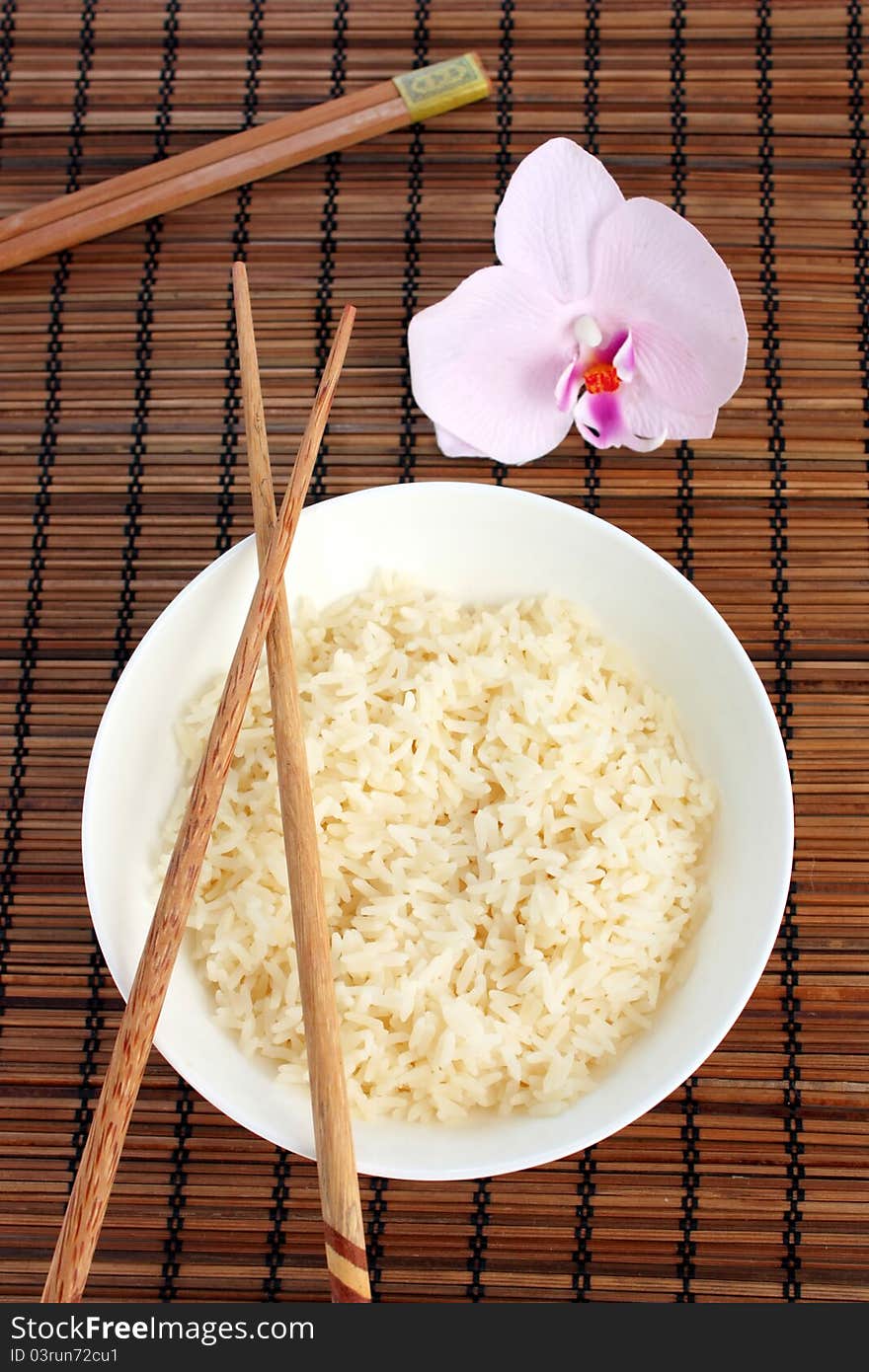 An Asian place setting consisting of a bowl of white rice with chopsticks and a pretty orchid. View from above. An Asian place setting consisting of a bowl of white rice with chopsticks and a pretty orchid. View from above.