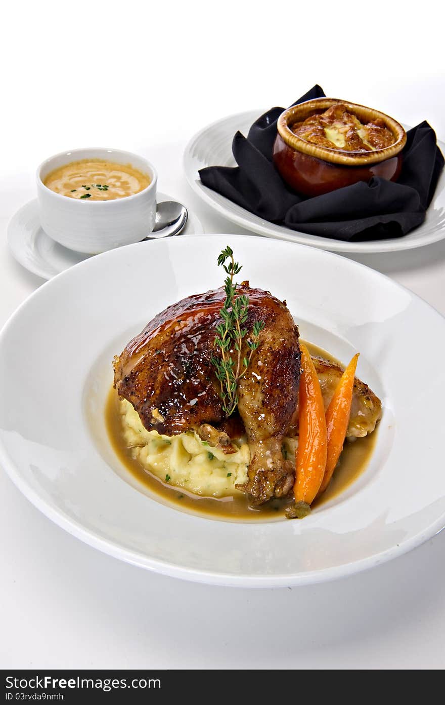 Rotisserie roasted chicken leg served over mashed potatoes with baby carrots and a gravy sauce. A fresh green sprig of rosemary tops this off. Warm bowls of soup are featured in the background. Rotisserie roasted chicken leg served over mashed potatoes with baby carrots and a gravy sauce. A fresh green sprig of rosemary tops this off. Warm bowls of soup are featured in the background.