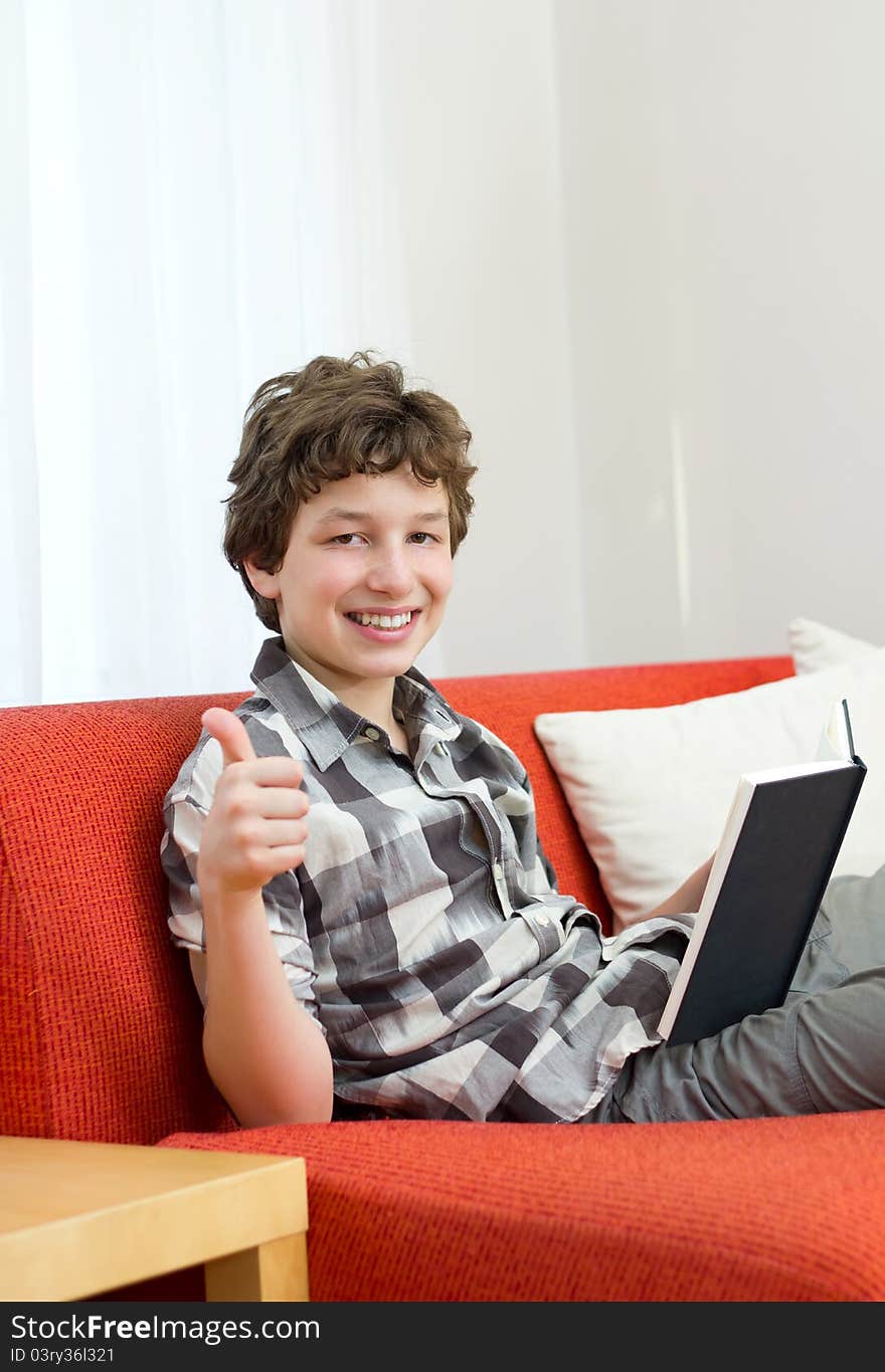 A preteen boy seated on an orange couch with white pillows gives a thumbs up and a big smile as he holds the book he was reading with his other hand. A preteen boy seated on an orange couch with white pillows gives a thumbs up and a big smile as he holds the book he was reading with his other hand.