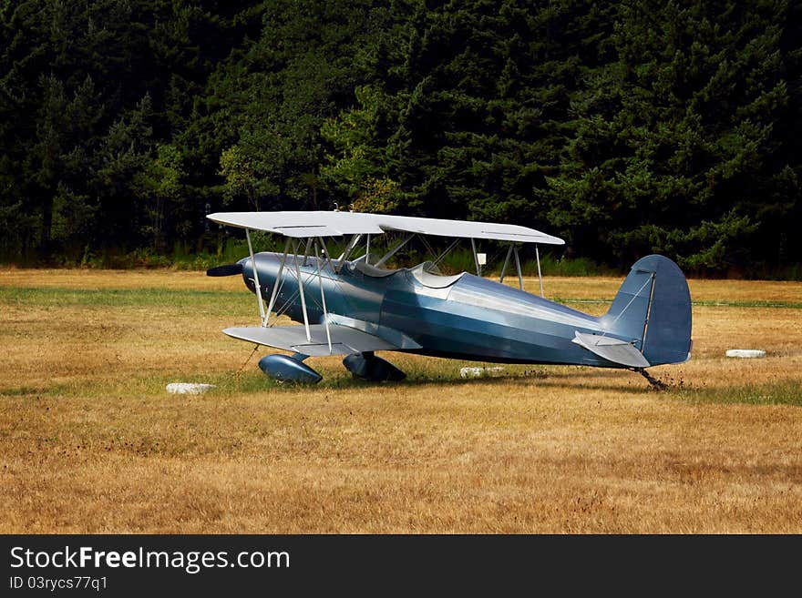 A beautiful blue Pitts S-2 Special biplane sits parked and tied down in a field with evergreen trees in the background. A beautiful blue Pitts S-2 Special biplane sits parked and tied down in a field with evergreen trees in the background.