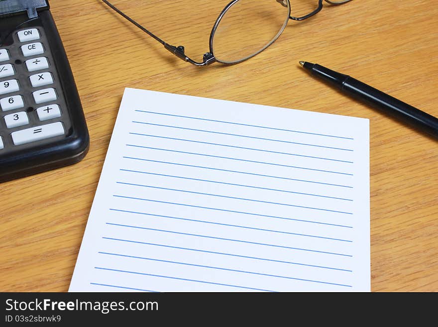 A blank lined pad on a wood desk with a calculator, eyeglasses, and pen. A blank lined pad on a wood desk with a calculator, eyeglasses, and pen