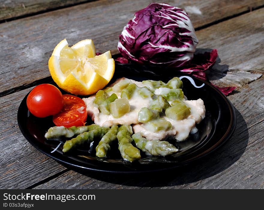 Veal escalope in black dish with asparagus, tomatoes and lemon on wooden table. Veal escalope in black dish with asparagus, tomatoes and lemon on wooden table