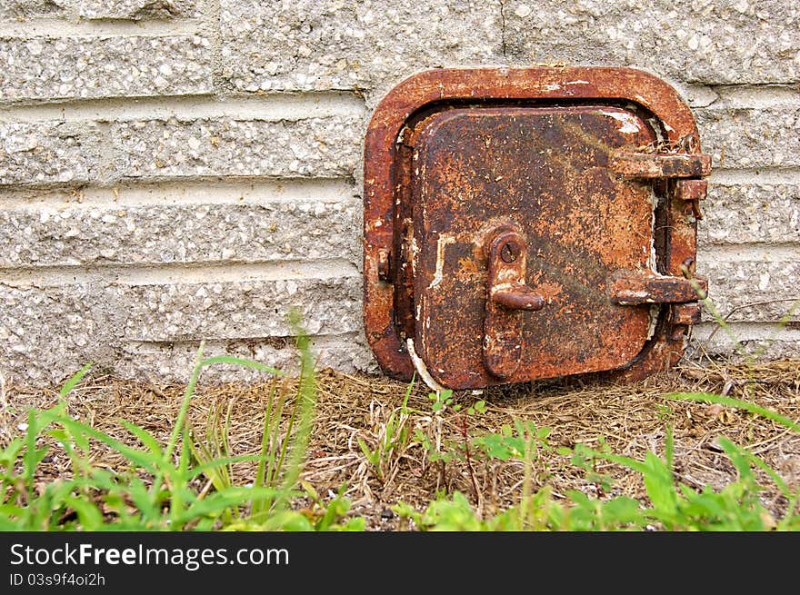 An old rusted iron door sits close to the ground on the outside wall of a stone brick chimney.