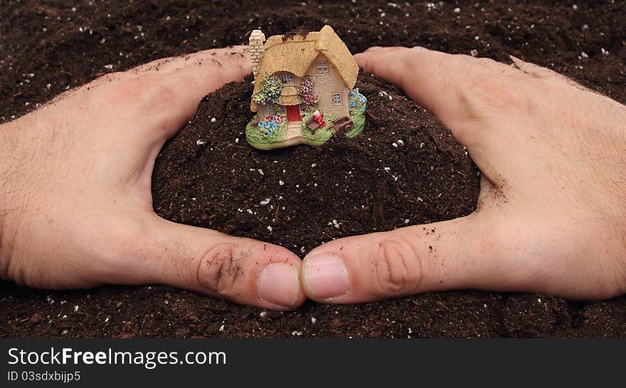 Two hands on brown soil making a circle with small country house in center. Two hands on brown soil making a circle with small country house in center