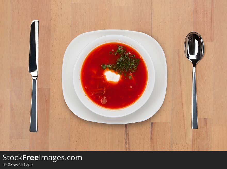 A plate of traditional Russian red borsch with sour cream and greens on a wooden table with spoon and knife.