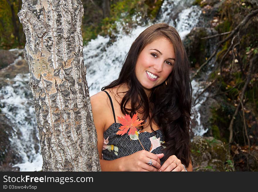 Young attractive smiling girl leaning up against a tree while holding a fallen leaf in autumn with a waterfall behind her. Young attractive smiling girl leaning up against a tree while holding a fallen leaf in autumn with a waterfall behind her