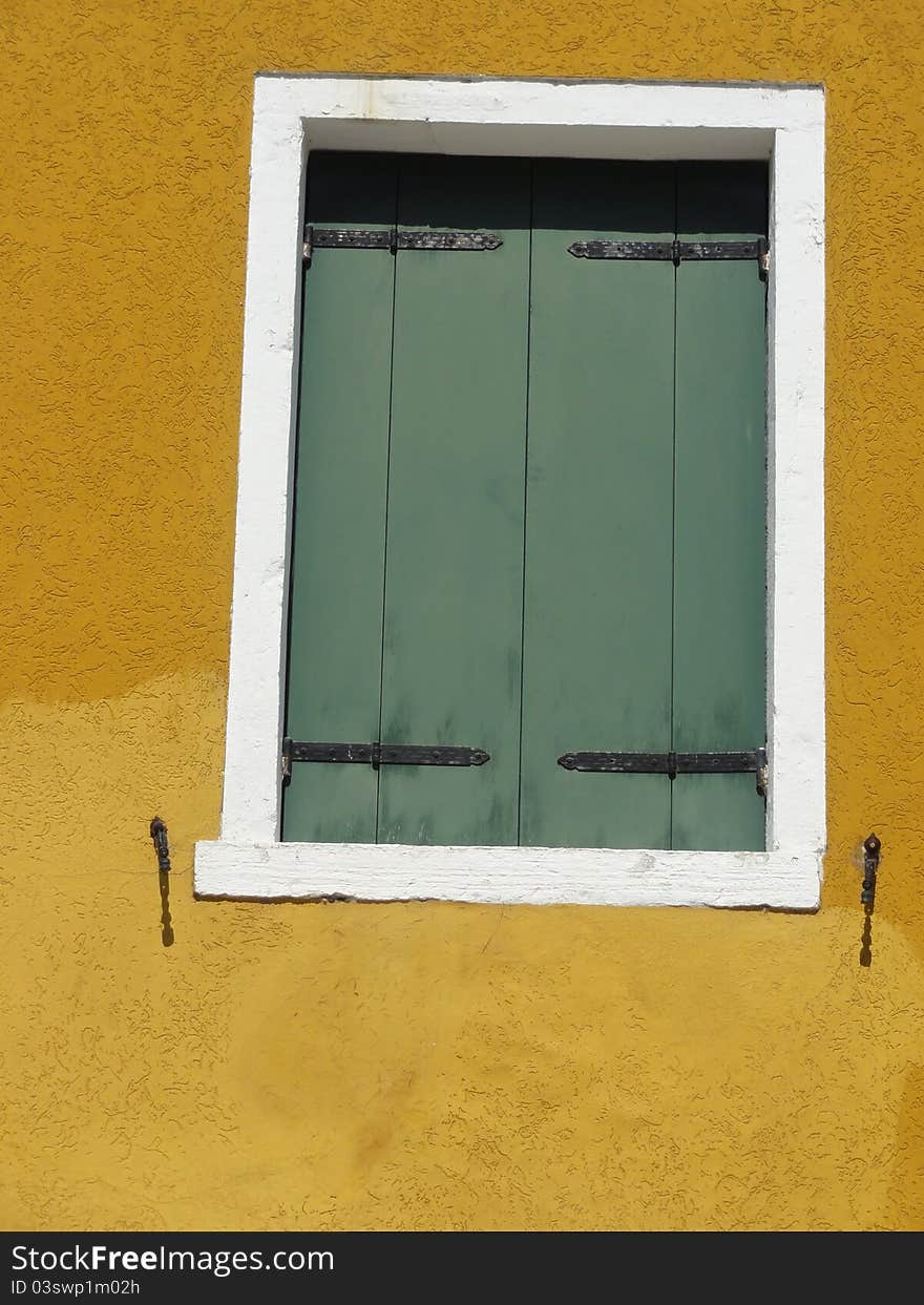 A green shuttered window, bordered by a white frame, in a yellow colored wall. A green shuttered window, bordered by a white frame, in a yellow colored wall.