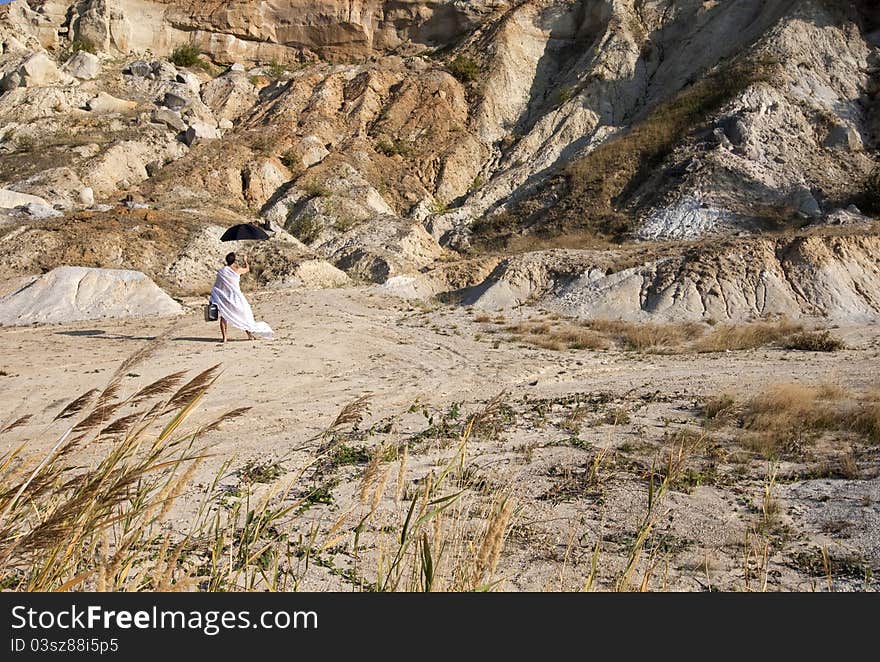 Man in white gown with a black suitcase and black umbrella walking in deserted land