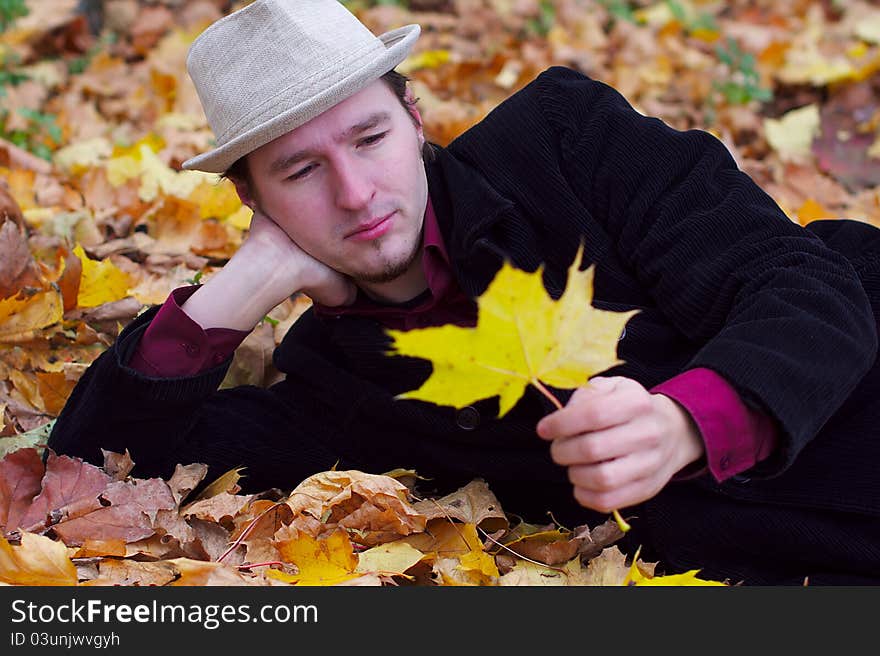 Handsome man with hat lying in autumn leaves and holding one leave. Handsome man with hat lying in autumn leaves and holding one leave