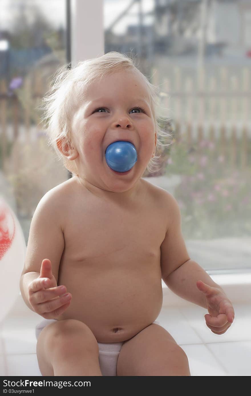 The little girl the blond, one year, sits on a white window sill, Plays with a dark blue ball and laughs. The little girl the blond, one year, sits on a white window sill, Plays with a dark blue ball and laughs